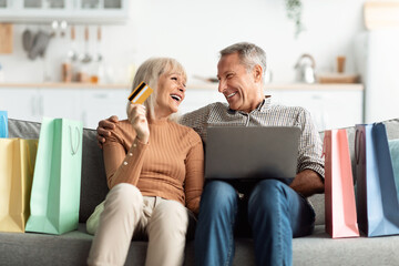 Obraz premium A joyful couple sits together on a couch surrounded by colorful shopping bags. They share smiles while holding a credit card and using a laptop for online purchases.