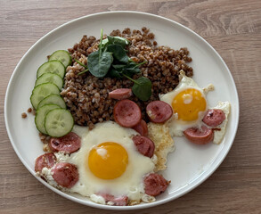 A hearty breakfast featuring fried eggs, sausages, buckwheat, and fresh cucumber slices on a plate