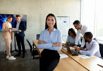 A beautiful asian businesswoman stands confidently with arms crossed in a stylish boardroom. She smiles at the camera while her colleagues engage in discussions nearby.