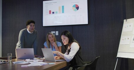 Discussing three-person team in office wear reviewing charts on monitor and laptops in meeting room