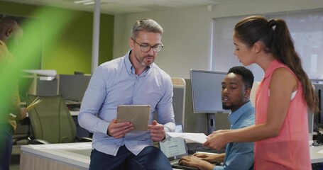 Speaking mature man sitting on desk edge holding tablet at open-plan office, woman passing paper
