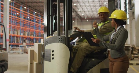 Directing female in blazer pointing clipboard to forklift driver in warehouse with yellow hard hats