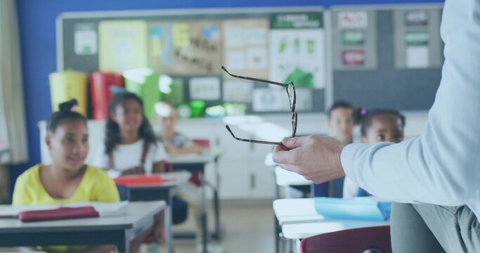 Holding eyeglasses, teacher in light shirt gesturing at front of class, pupils at desks, copy space