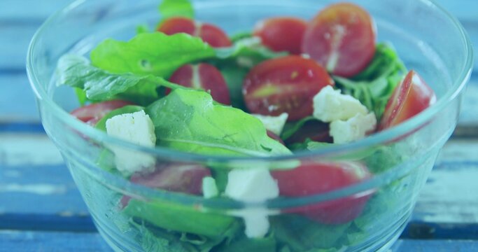 Showing clear glass salad bowl on painted blue table, with arugula, halved tomatoes, crumbled feta
