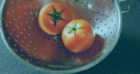 Showing perforated steel colander holding two ripe red tomatoes on kitchen countertop with droplets