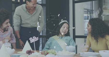 Smiling team gathering seated woman wearing blue top, sash, paper hat in meeting room, BABY sign
