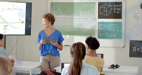 Holding tablet female teacher in blue top addressing class at front, with monitor, student laptops