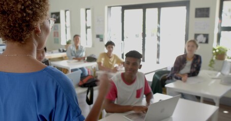 Teaching teacher blue top, glasses gesturing front-left at classroom with silver laptop, copy space