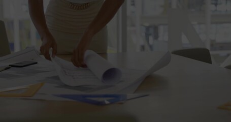 Standing in ribbed sleeveless dress handling blueprints on wood table near windows, blue set-square