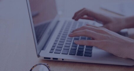 Typing woman's hands pressing keys on laptop keyboard at wooden desk, with eyeglasses nearby