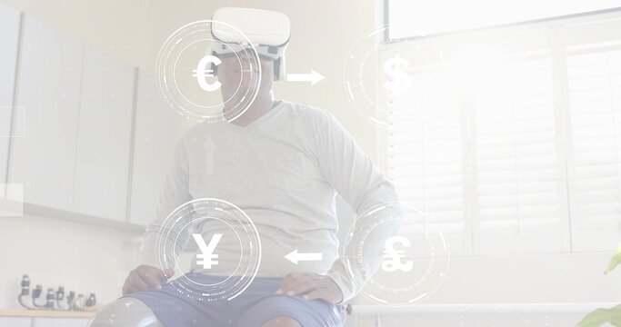 Sitting mature man wearing VR headset viewing currency icons in bright kitchen, near counter