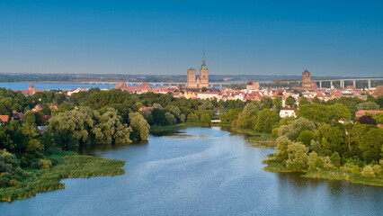 Obraz premium Panoramic aerial view of Stralsund, Germany, with historic brick churches, old town roofs and lush green trees reflected in tranquil ponds on a clear summer evening under deep blue sky.