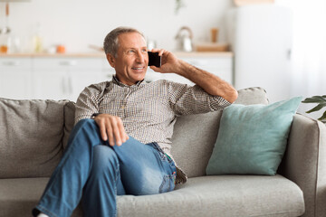 An elderly man smiles as he talks on his smartphone, seated comfortably on a soft couch in a well-lit living room during the afternoon.