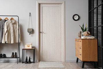 Interior of modern hall with beige door, chest of drawers and rack with clothes near grey wall