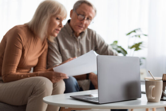 A senior couple sits closely on a couch, looking at documents while having a laptop open on a coffee table. Soft natural light fills the room, creating a cozy atmosphere.