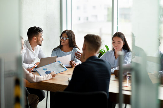 A cheerful group of young professionals from diverse backgrounds brainstorms during a corporate meeting. They engage in discussion about company strategy and success, sharing ideas in a bright office.