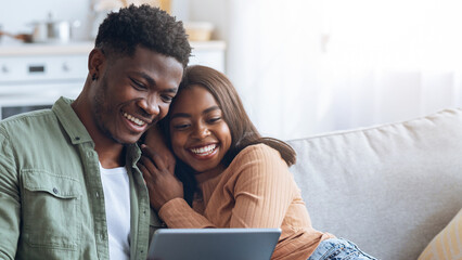 Two people are sitting closely on a couch, smiling and looking at a tablet. The airy living room is filled with natural light, creating a warm and inviting atmosphere.