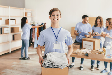 A cheerful young man holds a box of donated clothes in a donation center. Other volunteers are organizing donations and preparing for distribution. The atmosphere is friendly and collaborative.