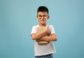 A cheerful boy with glasses embraces a stack of books against a light blue backdrop. His eagerness for learning and reading is clearly visible through his bright smile.
