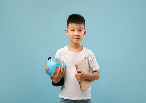 A cheerful young boy stands against a light blue background. He holds a globe in one hand and a notebook in the other, ready to explore. His bright smile adds to the scene's warm atmosphere.
