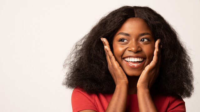 Joyful African American lady touches her face with excitement, looking to the side as if discovering an amazing offer. She stands against a clean white background, conveying happiness.