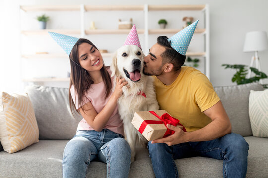 In a bright living room, a young couple celebrates a festive moment with their golden retriever. The woman smiles while the man kisses the dog on the head, holding a wrapped gift. - Powered by Adobe