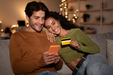 Happy couple sits on a cozy sofa at home in the evening, using a smartphone to shop online. They are smiling and holding a credit card, enjoying their purchasing experience.