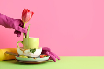 Female hand in rubber gloves with dirty dishes, detergent and tulip flower on color background