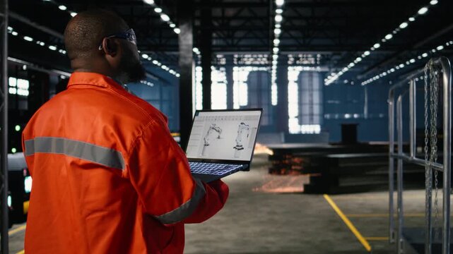 African american technician reviews robot software model on laptop in manufacturing plant, production tasks and equipment operation. Heavy industry with steel machinery, asset management. Camera A.