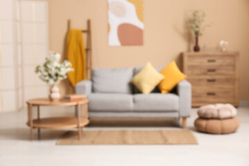 Blurred view of sofa with dresser, coffee table and vase of white blooming branches near beige wall in living room