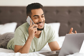 A cheerful young man talks on his phone while lying on a bed, accessing his laptop. The relaxed setting indicates a casual work or leisure moment, showcasing his engagement and focus.