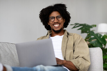 A cheerful young Black man with glasses happily uses his laptop while sitting on the couch at home. He enjoys his freelance work in a comfortable, bright living space with plants.