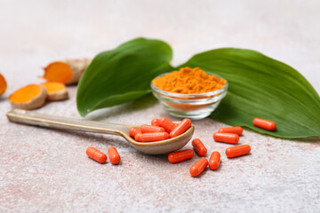 Spoon with capsules, leaves and bowl of turmeric powder on light beige grunge background, closeup