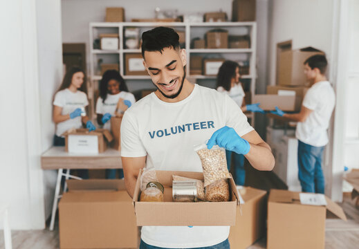 A man is smiling while holding a box of food items in a community center. Other volunteers are busy organizing supplies in the background. Everyone is wearing gloves and working together.
