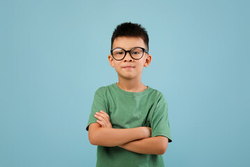 A young boy stands with his arms crossed, wearing glasses and a green shirt. He has a confident expression and is positioned in front of a light blue background, creating a cheerful vibe. © Prostock-studio