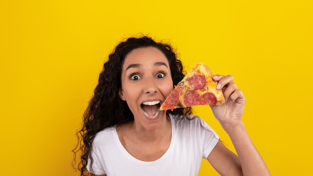 A woman with curly hair smiles widely while holding a slice of pepperoni pizza in front of a bright yellow backdrop. Her excitement suggests she loves the pizza.