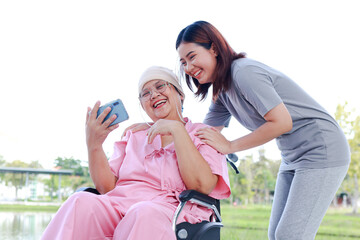 Caregivers or staff take care of elderly cancer patients wearing head coverings sitting in a wheelchair outside. They use smartphones to take pictures together. Daughter takes care of sick mother.