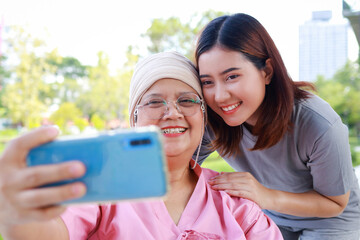 Caregivers or staff take care of elderly cancer patients wearing head coverings sitting in a wheelchair outside. They use smartphones to take pictures together. Daughter takes care of sick mother.