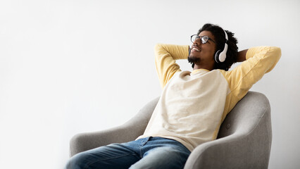 A young Black man is comfortably resting in a chair at home while wearing wireless headphones. He smiles as he enjoys his favorite music during a peaceful weekend.
