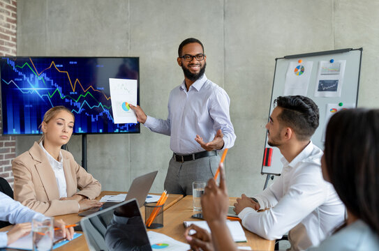 A confident African American boss stands in front of a screen, presenting a financial report to a diverse group of coworkers in a modern boardroom. Team members engage attentively during the meeting.