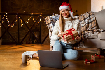 A happy woman in a Santa hat holds a beautifully wrapped gift while sitting on the floor. A laptop in front of her suggests a virtual holiday celebration. Festive lights create a warm atmosphere.