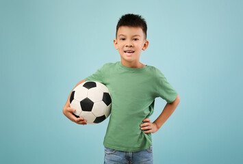A young boy with short black hair is smiling while holding a soccer ball. He wears a green shirt and denim jeans, standing against a light blue background.