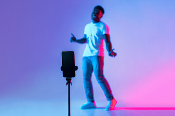 A young man shows off his dance moves in front of a smartphone placed on a tripod. The vibrant background features shades of blue and pink, creating a lively atmosphere.