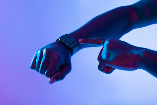 A hand fingers a smartwatch on the wrist, highlighted by vibrant blue and purple lighting. The scene conveys a sense of tech-savvy lifestyle and modernity.