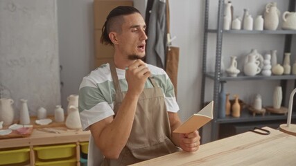 Man in apron writes in notebook holding pen and tapping chin while seated at pottery worktable with ceramic vases on shelves in studio; quiet contemplation.