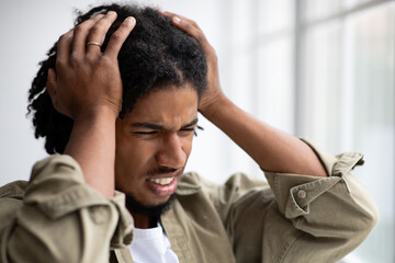 A young African American man is standing by a big window, visibly stressed and anxious. He is touching his head, showing signs of discomfort and worry, likely due to a headache.