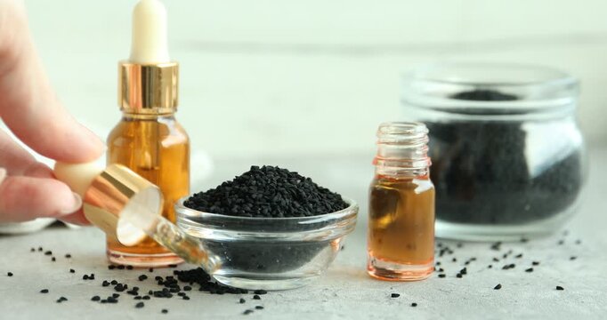 Woman taking pipette from bottle of essential oil and putting it onto grey table with black cumin seeds, closeup