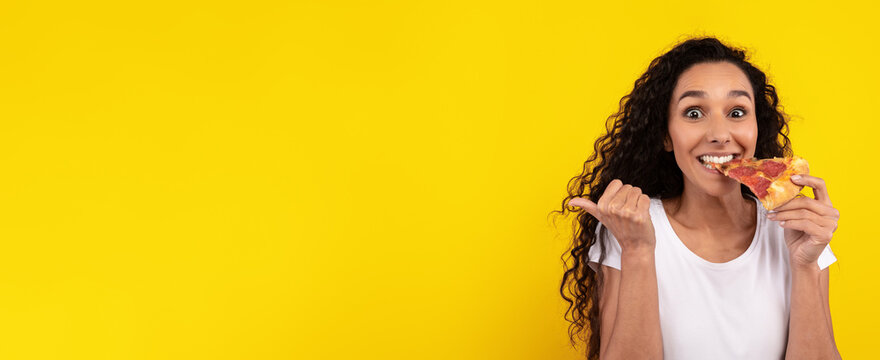 A woman with curly hair is smiling widely while holding a slice of pizza. She is standing in front of a vibrant yellow wall, clearly enjoying her snack.