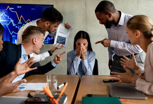 A group of aggressive coworkers shouts at a distressed Asian female employee during a corporate meeting. She covers her face in despair while facing multiple accusations in a boardroom.