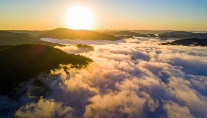 An aerial view of mountains and clouds at sunrise. The sun is shining brightly, casting a golden glow over the landscape.